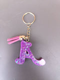 Porte-clé lettre A sculptée en résine violet améthyste à grosses paillettes holographiques irisées avec pompon suédine rose fuchsia et anneau doré