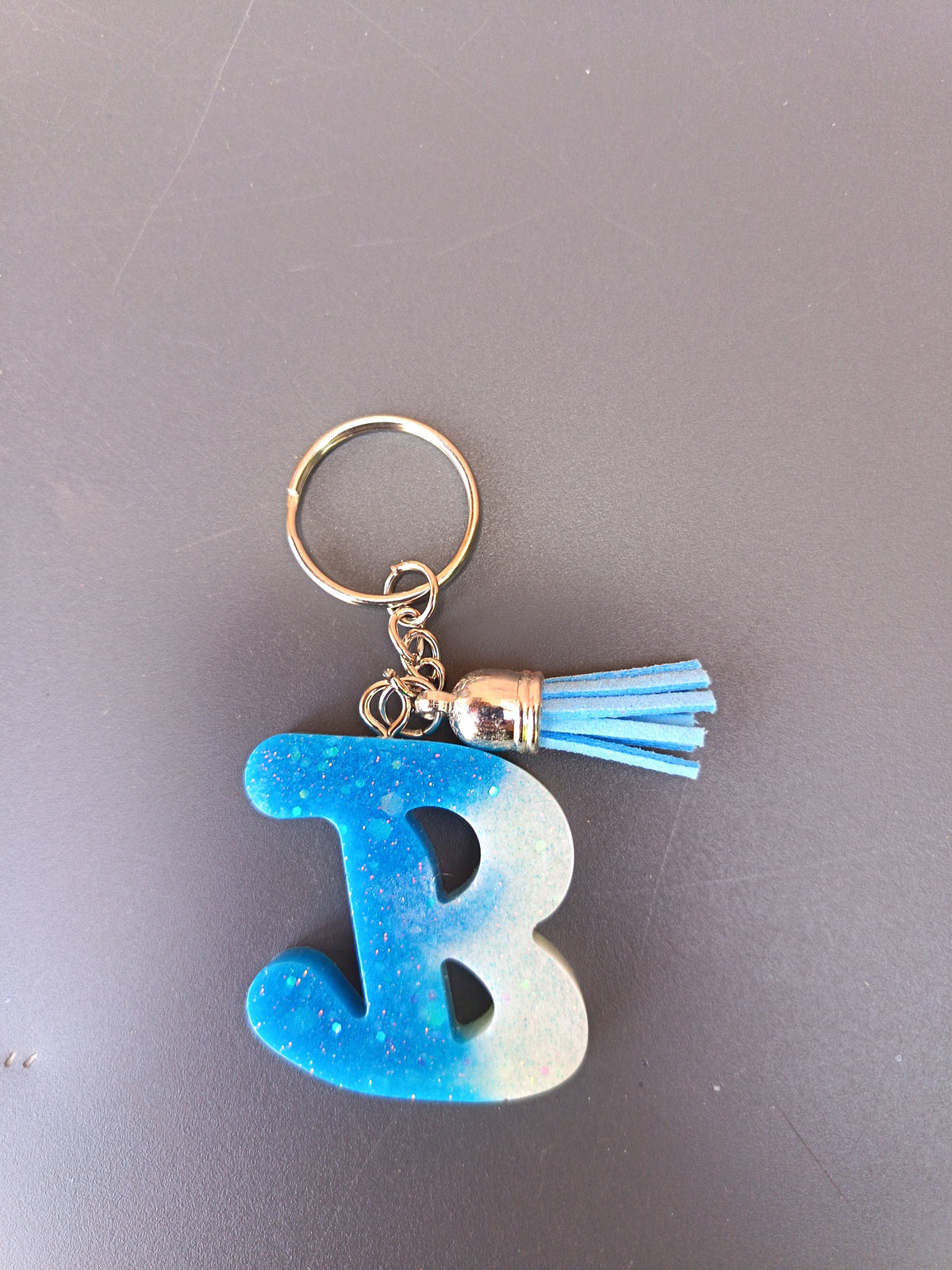 Porte-clé lettre B sculptée en résine dégradé bleu ciel et blanc nacré à micro-paillettes avec pompon suédine bleu ciel et chaînette dorée