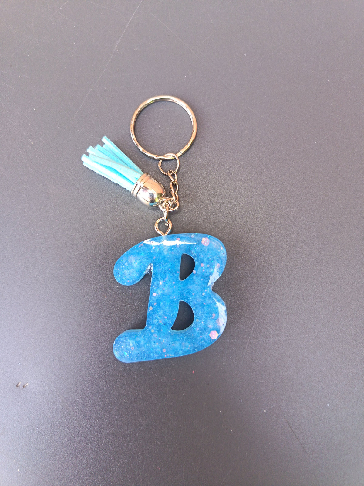 Porte-clé lettre B sculptée en résine bleu électrique translucide à grosses paillettes irisées roses et blanches avec pompon suédine bleu turquoise clair