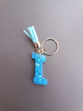Porte-clé lettre I sculptée en résine bleu turquoise à grosses paillettes holographiques avec pompon suédine bleu ciel et chaînette argentée